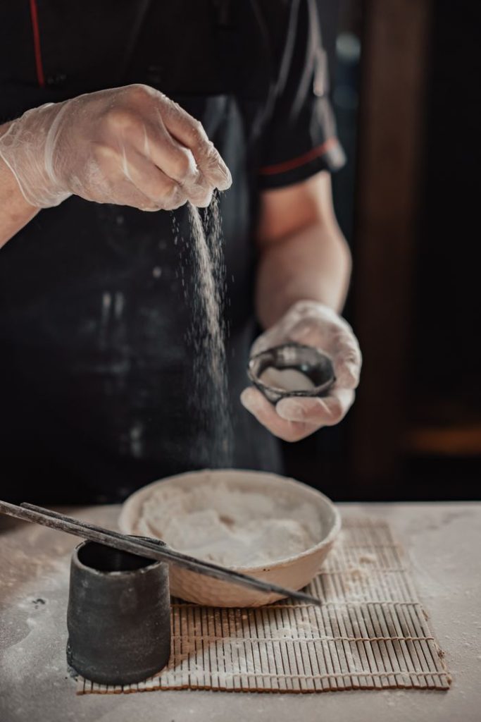 Close-up of a chefs hands sprinkling flour over a bowl of dough in a cozy kitchen setting.