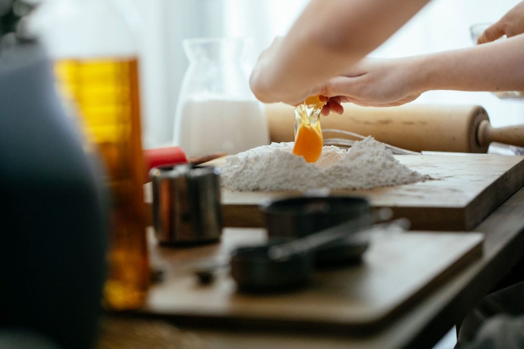 Unrecognizable cook breaking egg into pile of flour on wooden board while preparing dough at table in kitchen against blurred background